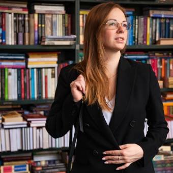 An adult woman stands in a library surrounded by shelves filled with books. She appears engaged and thoughtful, reflecting on her learning and development while appreciating the environment.